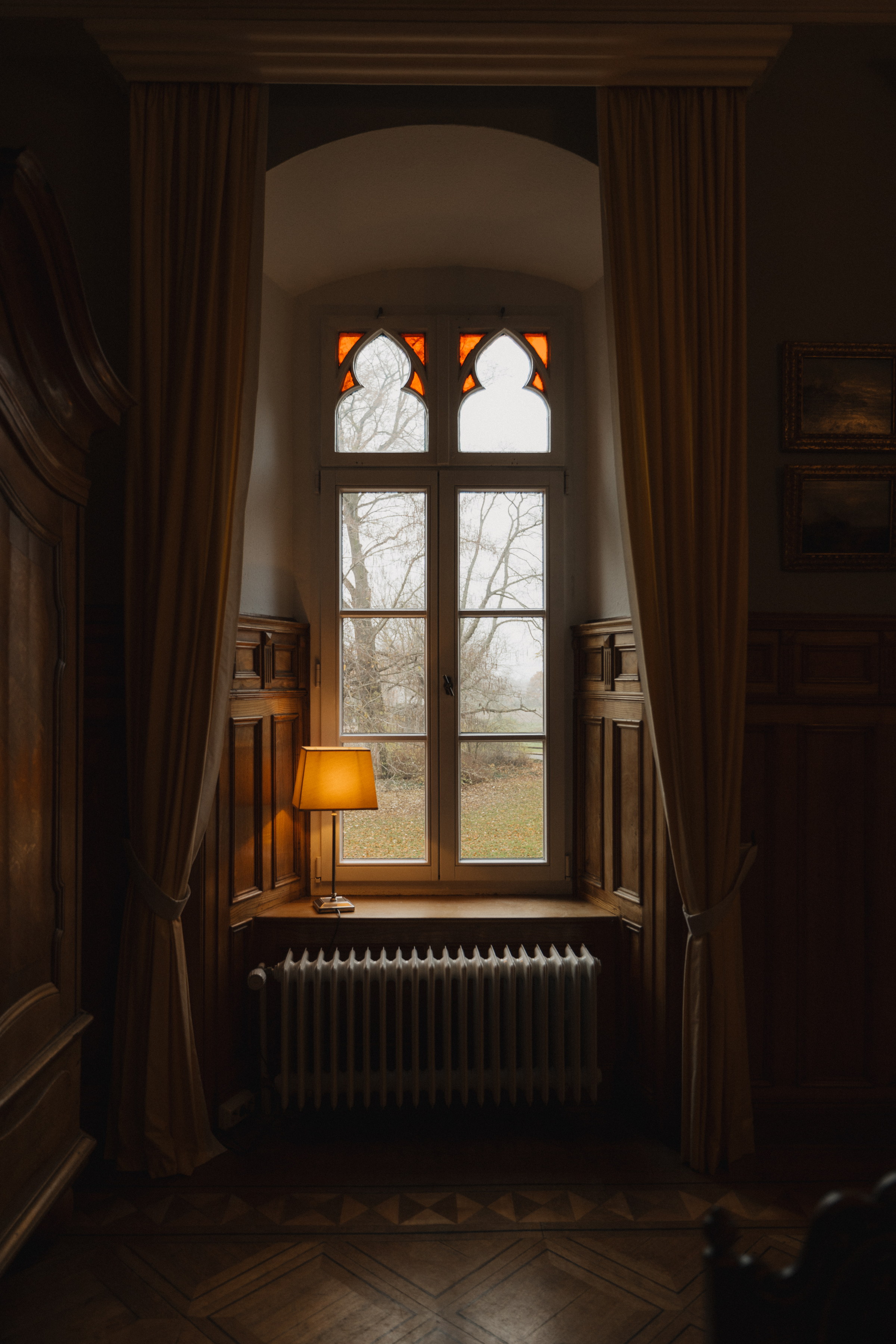 Spitzbogenfenster mit Tischlampe und Heizkörper im holzvertäfelten Raum der Burg