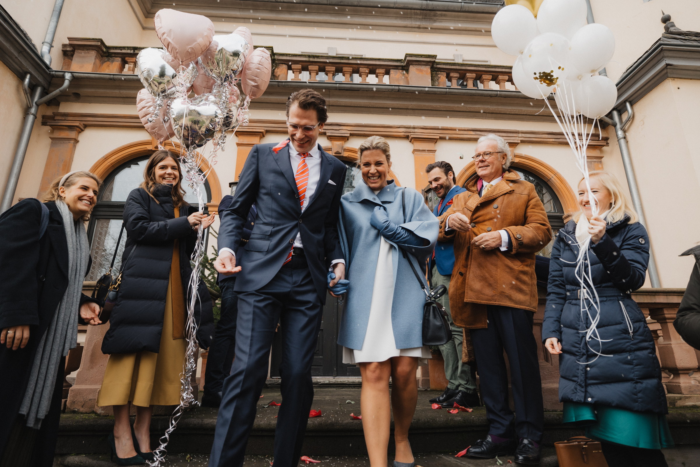 Brautpaar auf der Freitreppe zwischen Gästen mit Luftballons und Konfetti