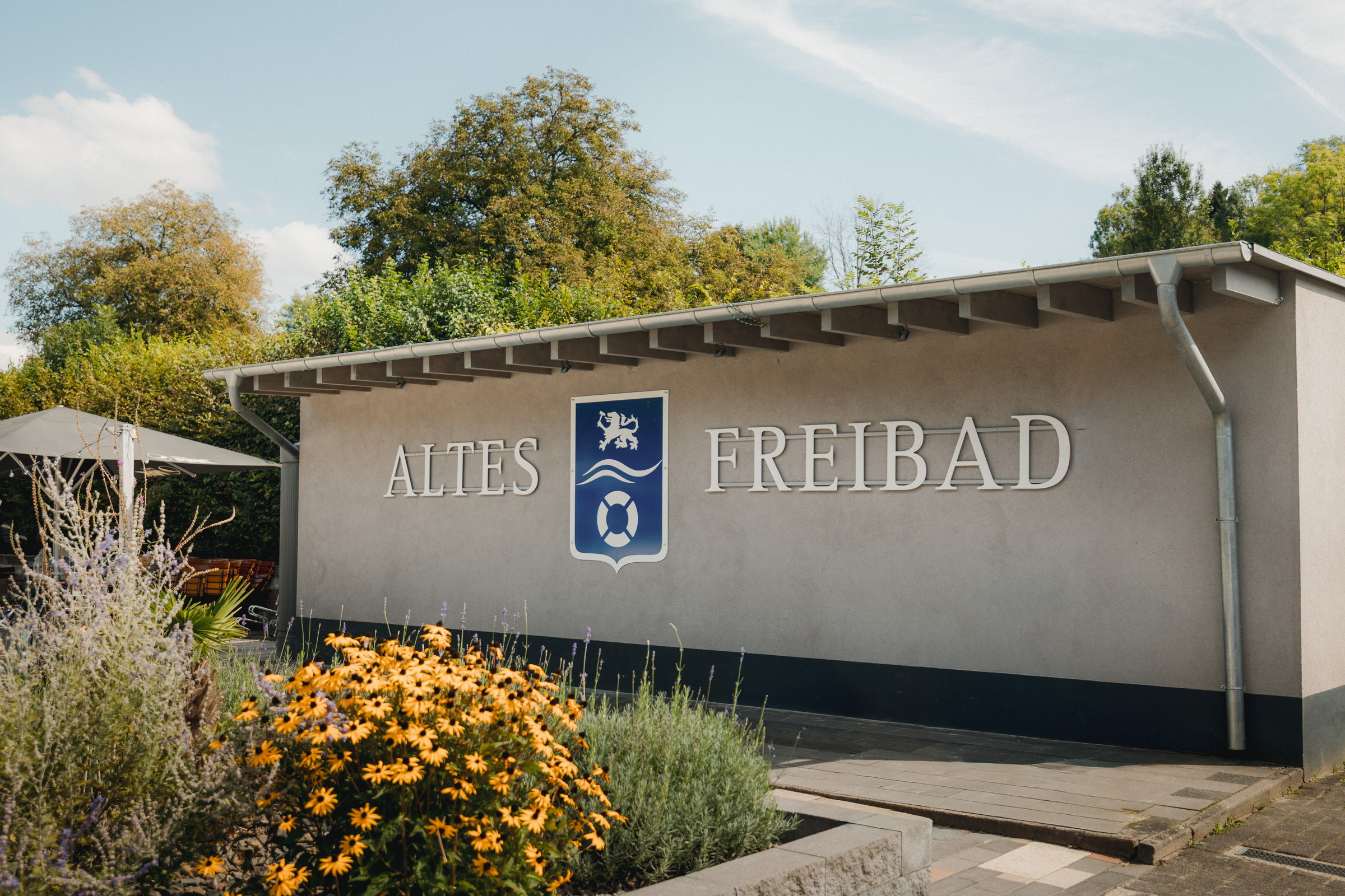 Eingangsgebäude mit dem Schriftzug Altes Freibad, davor ein Beet mit gelben Blumen