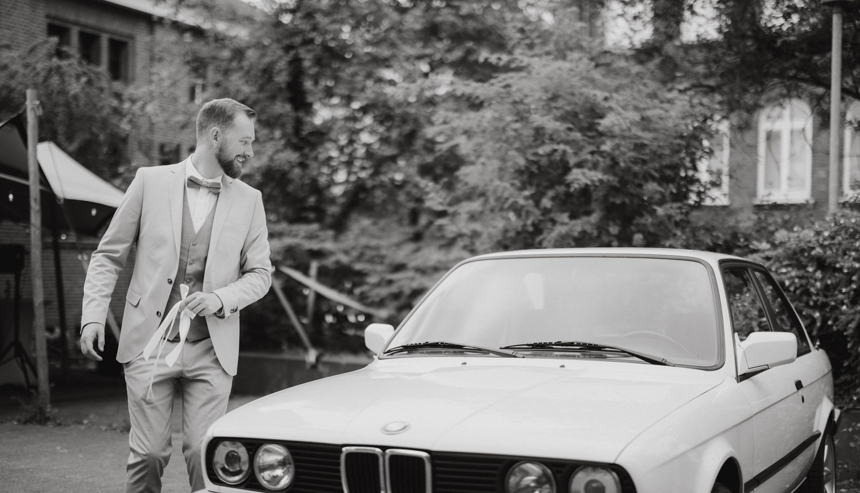 Bräutigam im hellen Anzug geht am weißen Oldtimer vorbei, Schwarzweiß