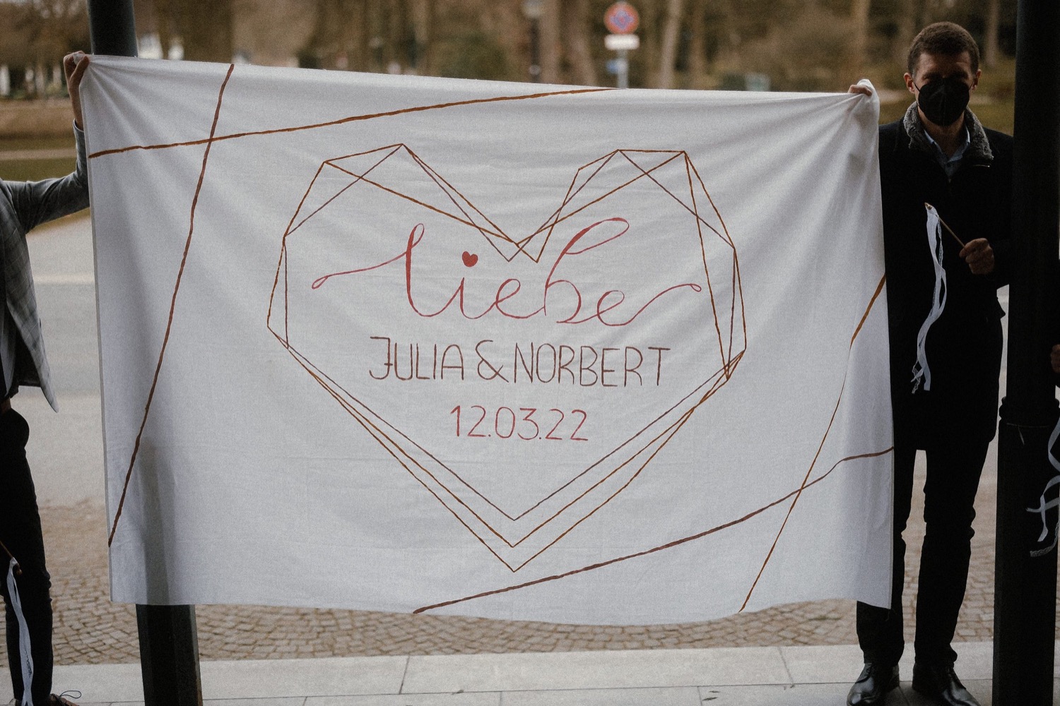 Gäste halten ein besticktes Herzbanner mit den Namen des Brautpaars und dem Hochzeitsdatum