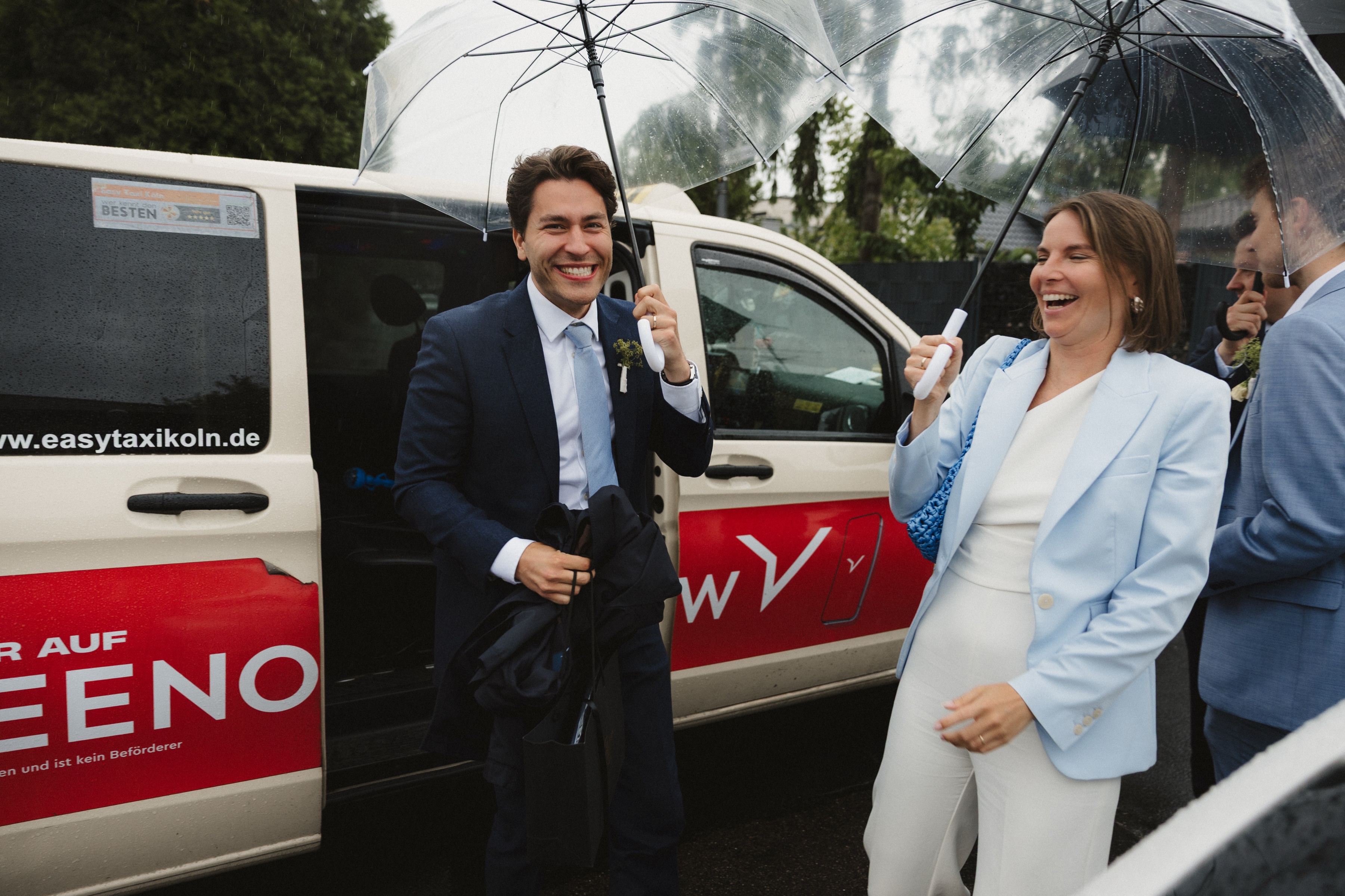 Brautpaar lacht unter Regenschirmen vor einem Taxi