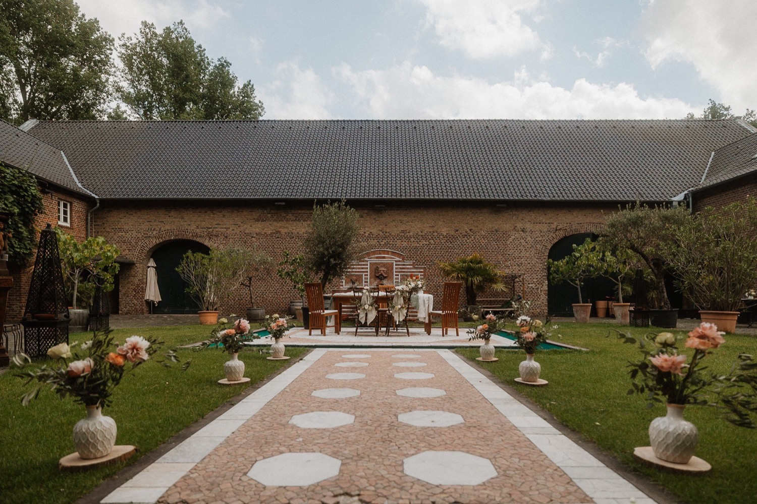 Innenhof mit langer Backsteinscheune, davor ein mit Blumenvasen gesäumter Weg zur Traubank