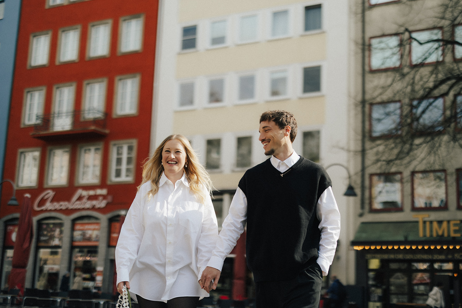 Paar geht lachend Hand in Hand über einen Platz in der Kölner Altstadt, dahinter ein rotes Haus