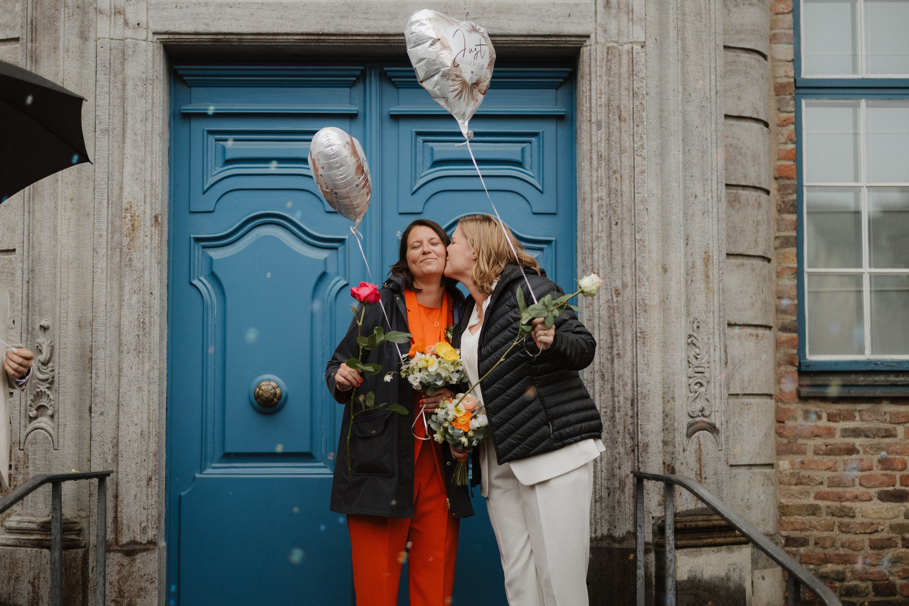 Das Paar küsst sich vor der blauen Rathaustür, mit Rosen und Just-Married-Ballons in den Händen