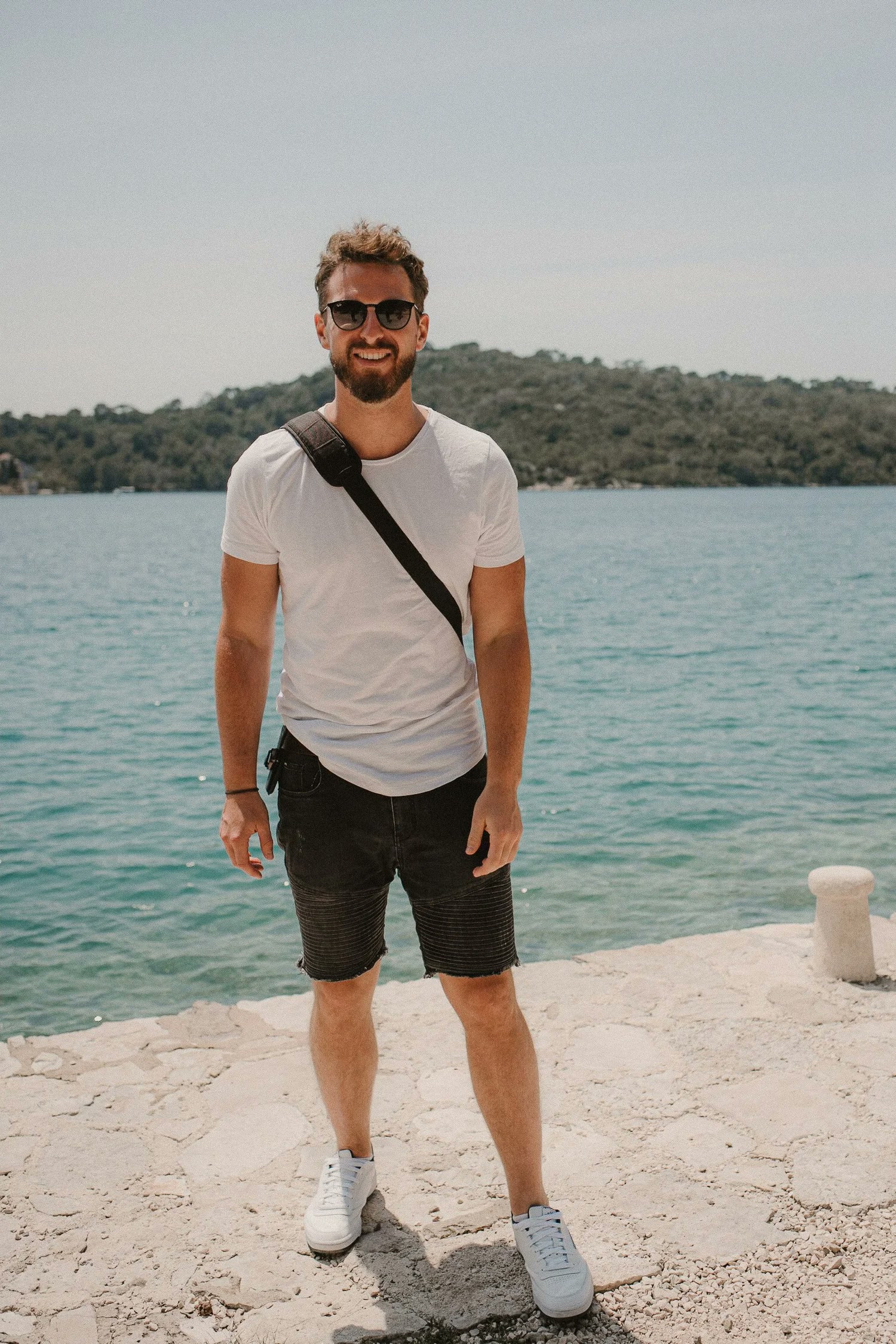 Fabian Rudolf mit Sonnenbrille und Umhängetasche an einer Bucht mit türkisem Wasser
