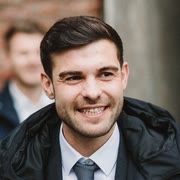 Mann in dunkler Jacke und Krawatte lächelt zur Seite